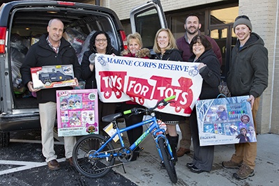 A group of seven adults pose in front of a van loaded with toys for the Marine Corps Reserve Toys for Tots charity drive. They are holding various donated toys including a bicycle, an LOL Surprise set, a toy police truck, and a Frozen II art set. The banner they display identifies this as a Marine Corps Reserve Toys for Tots donation event.