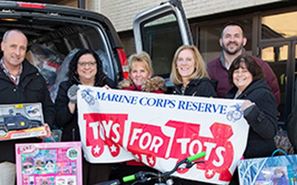A group of seven adults pose in front of a van loaded with toys for the Marine Corps Reserve Toys for Tots charity drive. They are holding various donated toys including a bicycle, an LOL Surprise set, a toy police truck, and a Frozen II art set. The banner they display identifies this as a Marine Corps Reserve Toys for Tots donation event.