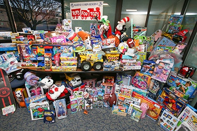 A large collection of donated toys is displayed on and around a table in front of a window, organized for the Marine Corps Reserve 'Toys for Tots' charity program. The toys span a wide variety of categories including stuffed animals, board games, action figures, vehicles, and dolls. The 'Toys for Tots' banner is prominently displayed at the top center of the image.