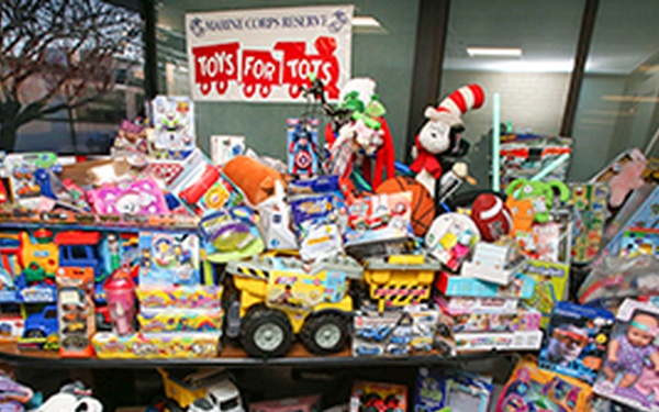 A large collection of donated toys is displayed on and around a table in front of a window, organized for the Marine Corps Reserve 'Toys for Tots' charity program. The toys span a wide variety of categories including stuffed animals, board games, action figures, vehicles, and dolls. The 'Toys for Tots' banner is prominently displayed at the top center of the image.