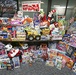 A large collection of donated toys is displayed on and around a table in front of a window, organized for the Marine Corps Reserve 'Toys for Tots' charity program. The toys span a wide variety of categories including stuffed animals, board games, action figures, vehicles, and dolls. The 'Toys for Tots' banner is prominently displayed at the top center of the image.