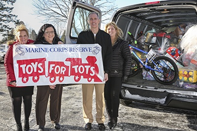 Four individuals are posing outdoors in front of a vehicle with its rear hatch open, holding a Marine Corps Reserve 'Toys for Tots' banner. The vehicle's cargo area is filled with donated toys, including a blue bicycle and various wrapped gifts and toy boxes. This appears to be a toy donation collection event for the Toys for Tots charitable program.