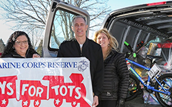 Four individuals are posing outdoors in front of a vehicle with its rear hatch open, holding a Marine Corps Reserve 'Toys for Tots' banner. The vehicle's cargo area is filled with donated toys, including a blue bicycle and various wrapped gifts and toy boxes. This appears to be a toy donation collection event for the Toys for Tots charitable program.