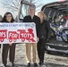 Four individuals are posing outdoors in front of a vehicle with its rear hatch open, holding a Marine Corps Reserve 'Toys for Tots' banner. The vehicle's cargo area is filled with donated toys, including a blue bicycle and various wrapped gifts and toy boxes. This appears to be a toy donation collection event for the Toys for Tots charitable program.
