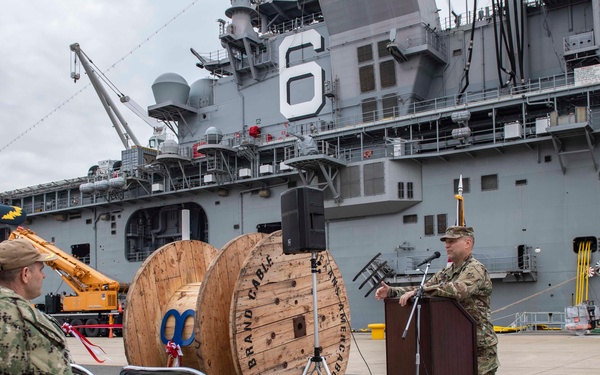 A military ceremony is taking place on a pier alongside a large naval vessel bearing the hull number '6'. A uniformed service member is speaking at a podium while other personnel in camouflage uniforms are seated as an audience. The Japanese national flag and a United States Navy flag are displayed, suggesting a joint U.S.-Japan naval event.