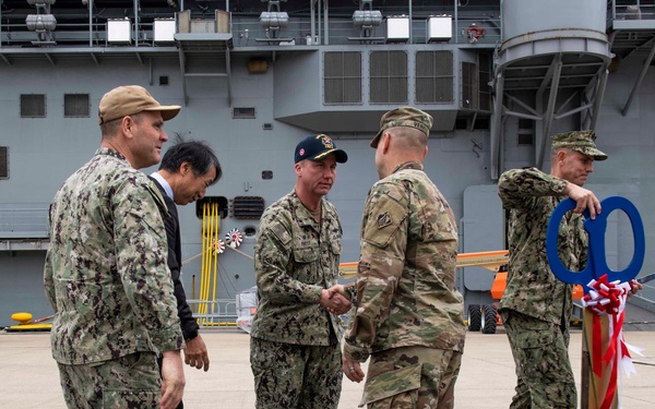 A group of military personnel in camouflage uniforms are gathered on what appears to be a naval vessel's flight deck or pier area, with a large gray warship visible in the background. Two individuals are shaking hands in the center while others observe, and one person on the right is holding large ceremonial scissors and a blue ribbon, suggesting a ribbon-cutting or commissioning ceremony. A civilian in a dark suit is also present among the uniformed personnel.