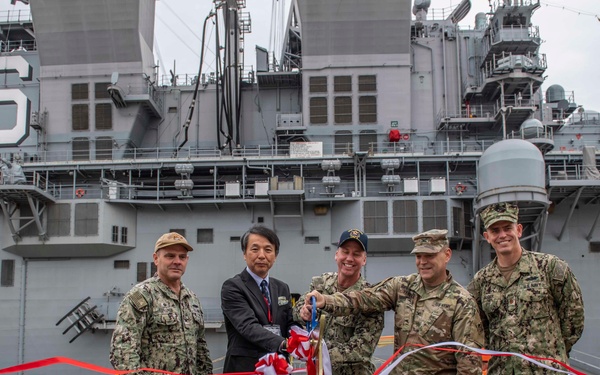 A ribbon-cutting ceremony is taking place in front of a large naval vessel bearing the hull number '6'. Five individuals — four in military camouflage uniforms and one in a civilian business suit — are jointly cutting red, white, and gold ceremonial ribbons. Large cable spools branded 'NEXANS AMERICAB' flank the group on both sides.