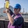 USS Wayne E. Meyer (DDG 108) Replenishment-at-Sea A young male worker wearing a blue hard hat and purple safety vest holds up a clear jar containing yellow liquid, likely inspecting a fuel or fluid sample. The setting appears to be the deck of a naval or military vessel during an underway replenishment operation, with a large supply ship visible in the background. The purple vest is consistent with U.S. Navy fuels personnel designation.