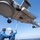 USS WAYNE E. MEYER (DDG 108) Flight Operations Two naval deck crew members in blue flight deck suits are using a pole to guide or secure a military helicopter during landing operations on what appears to be a naval vessel at sea. The image is taken from a dramatic low-angle perspective directly beneath the hovering aircraft, revealing its underside including landing gear, sensors, and external stores. A smaller unmanned aerial vehicle or drone is visible attached to or near the helicopter.