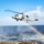 USS WAYNE E. MEYER (DDG 108) Flight Operations A U.S. Navy military helicopter, marked with the number '43', is captured mid-flight lifting off from or hovering over a naval vessel's flight deck above open ocean. The rotor wash is creating a mist spray over the water, which is refracting sunlight to produce a visible rainbow below the aircraft. The sky is mostly clear blue with scattered clouds in the background.