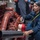 USS WAYNE E. MEYER (DDG 108) Shipboard Operations A person in navy blue coveralls is working in what appears to be a ship's mechanical or engine room, handling a red fire extinguisher near a complex system of red pipes and valves. They are wearing ear protection (orange earplugs) and have a yellow strap across their chest, suggesting safety compliance. Additional personnel are visible in the background observing or supervising.
