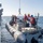 USS WAYNE E. MEYER (DDG 108) Shipboard Operations A rigid-hull inflatable boat (RHIB) named 'Brian Hicks' is being hoisted or lowered alongside a naval warship using a crane/davit system. Several personnel wearing hard hats and orange life vests are aboard the RHIB, while additional crew members observe from the ship's deck. The operation appears to be a standard small boat recovery or deployment exercise at sea.