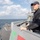USS WAYNE E. MEYER (DDG 108) Shipboard Operations A naval servicemember in dark uniform and cap stands watch on the bridge wing of a military warship at sea. The vessel appears to be a U.S. Navy guided-missile destroyer or similar surface combatant, with a helicopter landing pad visible on the aft deck. The ship is underway in calm, open ocean conditions under a partly cloudy sky.