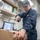 USS WAYNE E. MEYER (DDG 108) Shipboard Operations A U.S. Navy sailor in blue coveralls is scanning a cardboard package with a handheld barcode scanner in what appears to be a shipboard mail or logistics office. The sailor is wearing a cap with 'USS Wayne E. Meyer 108' insignia, indicating this is aboard a U.S. Navy destroyer. The workspace contains binders, a computer terminal, a telephone, and mail safety posters.