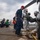 USS Wayne E. Meyer (DDG 108) Aviation Fire Fighting Drill Military personnel are conducting what appears to be a helicopter deck operations drill or exercise aboard a naval vessel. Two crew members in silver proximity/fire-resistant suits with yellow boots are assisting someone exiting or entering a helicopter, while other personnel in green flight deck gear crouch nearby. A supervisor in navy blue coveralls and a red cap oversees the activity on the flight deck.