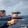 USS Wayne E. Meyer (DDG 108) Live Fire Gunnery Exercise Multiple personnel in blue uniforms are conducting a pistol qualification or training exercise on the deck of a naval vessel at sea. They are firing semi-automatic pistols over the ship's railing toward the open ocean. All visible participants are wearing eye protection and ear protection, consistent with standard range safety protocols.