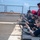USS Wayne E. Meyer (DDG 108) Live Fire Gunnery Exercise Multiple personnel are conducting a live-fire weapons qualification exercise on the deck of what appears to be a naval vessel at sea. Shooters are in the prone or low-supported position along a deck barrier, firing rifles downrange over the ship's side. A safety observer wearing a red vest and red hearing protection earmuffs stands behind the firing line.