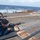 USS Wayne E. Meyer (DDG 108) Live Fire Gunnery Exercise Military personnel are conducting a small arms qualification or training exercise on the deck of a naval vessel at sea. A shooter in blue uniform is in the prone position aiming an M16/M4-style rifle, while an instructor in a red life vest provides guidance. Additional personnel with hearing protection are visible in the background near the ship's railing.