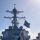 USS Wayne E. Meyer (DDG 108) Replenishment at Sea The image shows the forward section of a U.S. Navy Arleigh Burke-class guided-missile destroyer at sea, photographed from the bow looking aft toward the superstructure. A dark flag with the letters 'FOA' and a smaller yellow-green pennant are flying from the mast, suggesting a fleet operations or administrative signal. The ship's Mk 45 5-inch naval gun mount is prominently visible in the foreground.