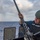 USS Wayne E. Meyer (DDG 108) Gun Shoot A U.S. Navy sailor in full combat gear is performing maintenance or inspection on a deck-mounted weapon system aboard a naval vessel at sea. The sailor is holding up what appears to be a barrel component of a large-caliber machine gun, likely conducting a barrel change or cleaning procedure. The setting is an open ocean environment with overcast skies and a faint rainbow visible in the background.