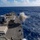 USS Wayne E. Meyer (DDG 108) Live Fire exercise A U.S. Navy warship, identifiable as an Arleigh Burke-class guided-missile destroyer, is firing its forward 5-inch (127mm) Mk 45 naval gun during what appears to be a live-fire exercise at sea. A large plume of gray-white gun smoke is visible at the muzzle, captured at the moment of discharge. The vessel is underway on open blue ocean under partly cloudy skies.