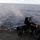USS Wayne E. Meyer (DDG 108) Gun Shoot A U.S. Navy sailor is operating a mounted machine gun on the deck of a naval vessel, firing into open ocean. Numerous spent brass shell casings are scattered across the deck, indicating sustained fire. A smoke plume is visible over the water where rounds are impacting, consistent with a live-fire training exercise.