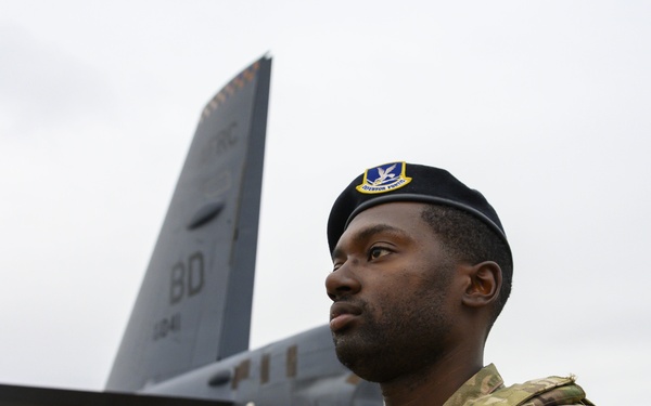 A U.S. Air Force Security Forces (SF) airman stands guard on a flight line, wearing multicam combat uniform and a black beret with a Security Forces badge. He is armed and positioned in front of a large military aircraft, identifiable by the tail code 'BD' and partial number '104'. The image appears to be an official military documentation or public affairs photograph.