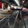 The image shows a military mess hall or galley serving line, photographed from a low angle at counter level. Food service workers wearing gloves are serving food onto blue trays for personnel in U.S. Navy digital camouflage (NWU) uniforms. The stainless steel steam table serving line contains multiple food items including what appears to be buffalo wings/chicken and other dishes.
