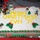 A large rectangular Christmas cake with white frosting is displayed on a red tablecloth. The cake is decorated with yellow icing text reading 'MERRY CHRISTMAS! CVN 71' and features fondant figurines including snowmen, penguins, an igloo, Santa Claus, Christmas trees, holly decorations, and gift boxes. The border is decorated with alternating red and green frosting piping.