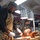 A U.S. Navy service member in camouflage uniform and apron is carving a roasted turkey on a wooden cutting board under heat lamps. The setting appears to be a military ship's galley or mess hall, likely during a holiday meal service such as Thanksgiving. Decorative elements and food displays are visible in the background.