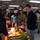 A U.S. Navy holiday meal event, likely a Christmas celebration, taking place in a military mess hall or galley aboard a ship or base. A cook in black culinary uniform is serving food to personnel and their families, including a child dressed in a Santa outfit. The setting features festively decorated serving tables with holiday greenery and food displays.
