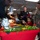 A holiday meal event aboard a U.S. Navy vessel, likely a Christmas celebration, where Navy personnel in NWU Type III camouflage uniforms are serving food to attendees including a young girl wearing a Santa hat and red dress. A decorated buffet table with a red tablecloth features elaborate food displays including carved vegetables, strawberries, and a roasted turkey or ham. The setting appears to be a ship's mess deck or galley.