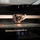 The image shows a close-up view of a 3D printer's print head assembly mounted on a horizontal aluminum extrusion rail. The hotend/extruder carriage is positioned near the glass print bed, with visible wiring and a small cooling fan attached. The print bed shows signs of previous use with residual filament material visible on the surface.