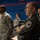 The image shows a group of military personnel in an indoor facility with blue walls. The foreground features a bald man in an olive/green flight suit with patches, standing with arms crossed, while others in US Air Force ABU (Airman Battle Uniform) camouflage and flight suits are visible in the background. The setting appears to be a military operations or briefing room with computer monitors visible.