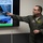 A military officer in an olive green flight suit is giving a briefing or presentation, pointing at a display screen showing images related to an F-22 aircraft. The screen displays text referencing 'ports/openings (F-22)' and is marked 'FOR OFFICIAL USE ONLY' at the bottom. The presenter appears to be explaining technical details about the aircraft's features.