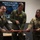 Three U.S. Air Force personnel are conducting a ribbon-cutting ceremony for the 1st Fighter Wing (1st FW) Innovation Lab. Two individuals in olive green flight suits and one in a MultiCam/OCP uniform are cutting a red ribbon with large ceremonial scissors. A display screen in the background reads 'Welcome to the 1st FW Innovation Lab!' with an image of a military aircraft.