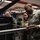 U.S. Air Force personnel are gathered in what appears to be a technical operations or training facility. A senior non-commissioned officer (identifiable by chevron rank insignia and 'U.S. Air Force' tape) is demonstrating or explaining equipment to a junior airman. Multiple uniformed personnel are visible in the background engaged in similar activities.