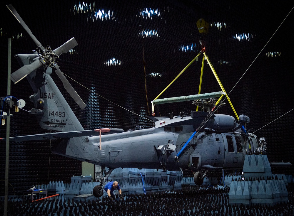 A USAF military helicopter, tail number 14483, is positioned inside a large anechoic chamber lined with pyramid-shaped sound/RF-absorbing foam panels on all surfaces. The aircraft appears to be suspended or supported by a yellow lifting rig attached to an overhead crane, suggesting maintenance, testing, or installation work. At least one technician in a blue shirt is working near the base of the aircraft.