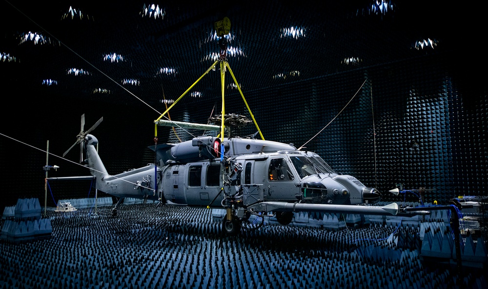 A military helicopter, consistent with a Sikorsky UH-60 Black Hawk or MH-60 variant, is positioned inside a large anechoic chamber for electromagnetic compatibility (EMC) or radio frequency (RF) testing. The chamber walls, floor, and ceiling are covered with pyramid-shaped RF-absorbing foam tiles to eliminate signal reflections. The helicopter is suspended and stabilized with yellow support rigging and cables connected to the ceiling structure.