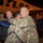 A nighttime scene on an airport tarmac showing military personnel in camouflage uniforms greeting each other with embraces near an aircraft. The partial text 'TLAS' is visible on the aircraft fuselage, consistent with Atlas Air livery. The atmosphere appears celebratory or welcoming, suggesting a homecoming or arrival event.