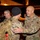 A nighttime military scene on what appears to be an airport tarmac where U.S. Army soldiers are greeting each other, likely during a deployment departure or return. The soldier on the right, wearing U.S. Army insignia and a unit patch, is embracing or greeting a soldier carrying a large tactical backpack. The warm, orange-tinted lighting suggests artificial airfield lighting in the background.