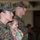 A female U.S. Marine in MARPAT digital camouflage uniform, identified by the name tape 'SZYPLK' (partially visible), stands at attention while holding a young child dressed in a floral outfit. Behind her stands a male Marine in the same uniform pattern. The setting appears to be an indoor military ceremony or formation with additional personnel visible in the blurred background.