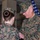 Two U.S. Marines in MARPAT woodland camouflage uniforms are engaged in what appears to be a military pinning or award ceremony. A senior male Marine is attaching or presenting something to the uniform of a female Marine whose back is to the camera. An American flag is visible in the background.
