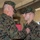 Two U.S. Marines in MARPAT woodland camouflage uniforms are engaged in a military award or decoration ceremony. An older, senior Marine is pinning a medal or decoration onto the chest of a younger Marine, who stands at attention. A red presentation box is visible, indicating a formal military commendation event.
