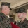 Two U.S. Marines in MARPAT woodland camouflage uniforms are engaged in what appears to be a military awards ceremony. The younger Marine on the right is wearing a medal with a green and orange ribbon and holding a red presentation folder, indicating he has just received a commendation or award. The older Marine on the left appears to be a senior officer or NCO conducting or concluding the presentation.