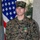 A U.S. Marine in MARPAT woodland digital camouflage uniform stands at attention in front of an American flag. The Marine, identified by name tape as 'WALLER,' is wearing a recently awarded medal on their chest. The setting appears to be a formal military award ceremony.