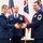 Three U.S. Air Force personnel in dress blue uniforms are participating in a formal military ceremony. Two individuals on the left and right are jointly presenting or receiving a wooden award/plaque box, while a third stands at attention in the center. The Ohio state flag and U.S. flag are visible in the background, suggesting this is an Ohio Air National Guard event.