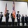 A military color guard ceremony is taking place on an indoor stage, with four uniformed personnel presenting flags including the United States flag and what appears to be the Ohio state flag. Three additional military personnel in dress uniforms stand at attention to the right side of the stage. The setting appears to be a military installation or armory facility.