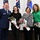 A U.S. Air Force senior non-commissioned officer in dress blues poses with three civilian women, each holding wrapped flower bouquets. The setting appears to be an official military ceremony or recognition event, with multiple flags visible in the background including the U.S. flag and what appears to be the Ohio state flag. The group is smiling, suggesting a celebratory or award-presentation occasion.
