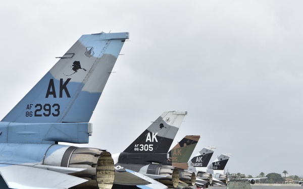 A row of U.S. Air Force F-16 Fighting Falcon jets are parked on a flight line ramp, displaying tail codes indicating Alaska Air National Guard assignment. The aircraft show varied camouflage schemes including air superiority blue-gray and aggressor-style paint. Ground support equipment is visible alongside the aircraft on what appears to be a tropical or warm-climate air base.